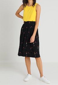 Top sin mangas amarillo combinado con una falda plisada negra que presenta un estampado floral en rojo y azul, complementado con zapatillas blancas.