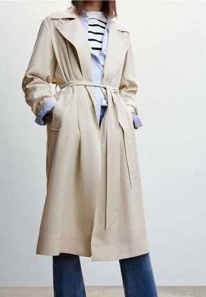 Persona con un abrigo trench beige con cinturón atado, sobre una camisa de rayas y vaqueros azules, de pie frente a un fondo claro y uniforme.
