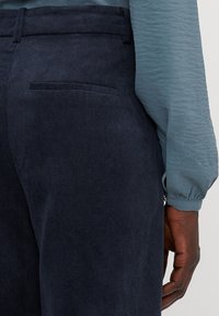 Navy corduroy pants with vertical ribbing, featuring a back pocket and a fitted waistband, paired with a textured light blue blouse.