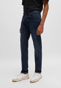 Sötétkék farmernadrág, slim-fit szabással, öt zsebbel, enyhe kopás részletezéssel, fehér sportcipővel párosítva.
