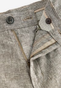 Pantalones de color marrón claro hechos de tela texturizada. Incluyen botones oscuros y cierre de cremallera. La cintura forrada presenta un patrón geométrico.