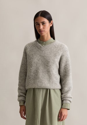 Gestrickter grauer Pullover mit V-Ausschnitt, der ein strukturiertes Muster aufweist. Darüber wird ein grün-weiß kariertes Kleid mit langen Ärmeln und einem Kragen getragen.