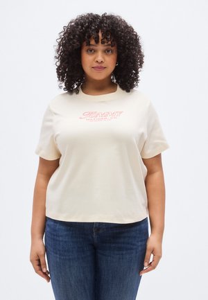 Frau mit lockigem Haar, die ein cremefarbenes Tommy Hilfiger T-Shirt und blaue Jeans trägt, steht vor einem schlichten weißen Hintergrund.
