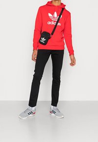 Röd huvtröja med vit logotyp, svarta tajta byxor och grå sneakers med blå och röda ränder. Liten svart axelremsväska.