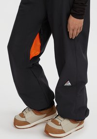 Zwarte broek met een oranje rits ventilatie in de dij, beige en bruine sneakers, en de hand van een persoon rustend aan de zijkant.