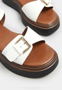 Sandali platform bianchi con una fascia larga, fibbia dorata e soletta in pelle marrone. Presentano una suola nera spessa per un'altezza aggiuntiva.
