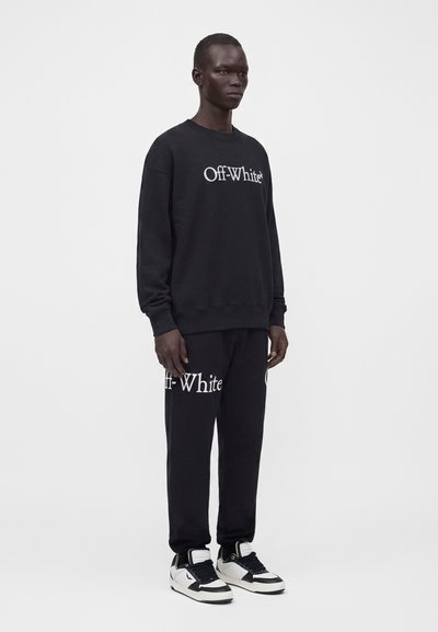 Άνδρας που φορά μαύρο φούτερ Off-White και αντίστοιχο παντελόνι φόρμας με λευκό κείμενο, φοράει μαύρα και λευκά αθλητικά παπούτσια μπροστά σε μονόχρωμο φόντο.