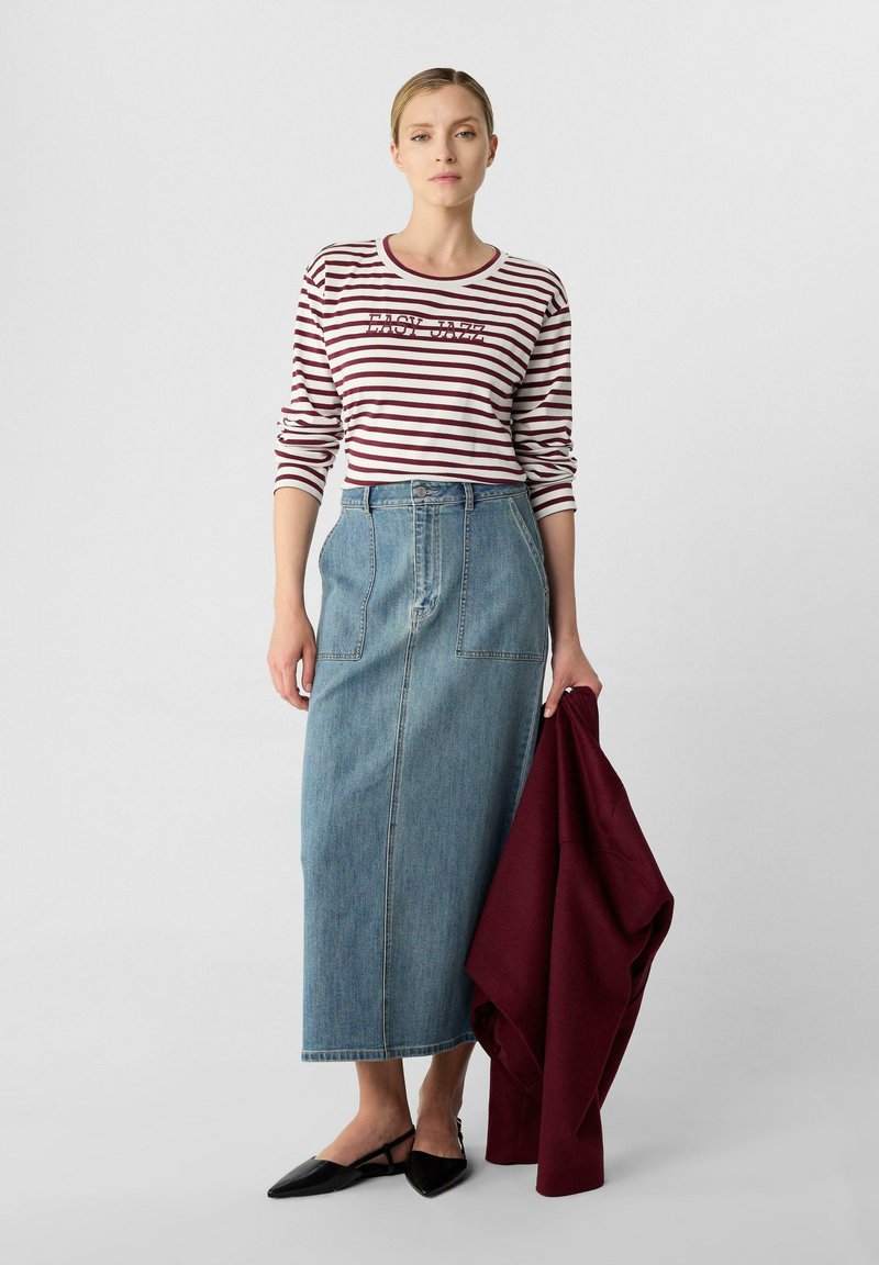Camicia a righe con maniche lunghe e scollo rotondo, abbinata a una gonna maxi in denim con tasche applicate. Ballerine nere a punta e giacca bordeaux.