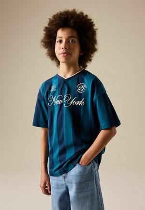 Dreng med krøllet hår iført en blåstribet atletiktrøje med "New York" og lyse denimjeans, stående med den ene hånd i lommen mod ensfarvet baggrund.
