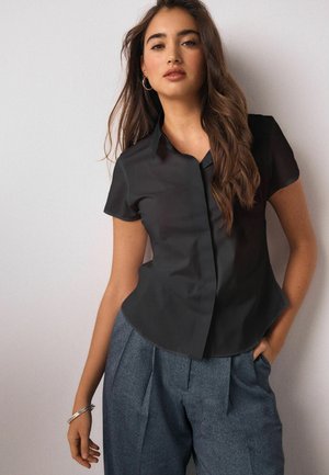 Sort kortærmet skjorte med knapper og krave, glat tekstur og tætsiddende facon. Kombineret med grå, plisserede bukser.