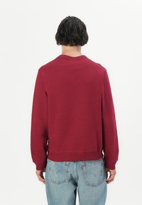 Burgundy sweatshirt with a crew neckline, ribbed cuffs, and hem. Paired with light blue denim jeans, visible back pocket detailing.