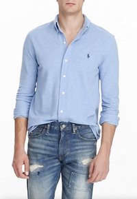 Lichtblauw overhemd met lange mouwen en een kraag, voorzien van een klein geborduurd logo op de borst en opgerolde mouwen, gecombineerd met vervaagde blauwe jeans.