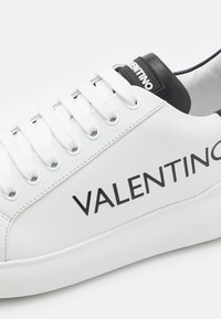 Bijele kožne tenisice s crnim detaljima, s ravnim bijelim vezicama, zaobljenim vrhom i istaknutim logotipom "VALENTINO" sa strane.