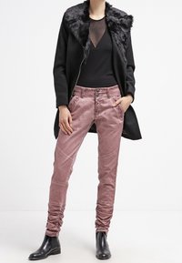 Svart kappa med fuskpälskrage, genomskinlig svart topp och ljusrosa byxor med texturerad detaljering. Svarta ankelboots fulländar outfiten.