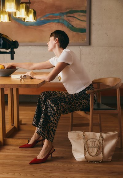 Weißes Kurzarmoberteil, weit geschnittene Leopardenmusterhose, rote High Heels, beigefarbene Tote-Bag mit Kettenriemen, Holztisch und Stuhl.