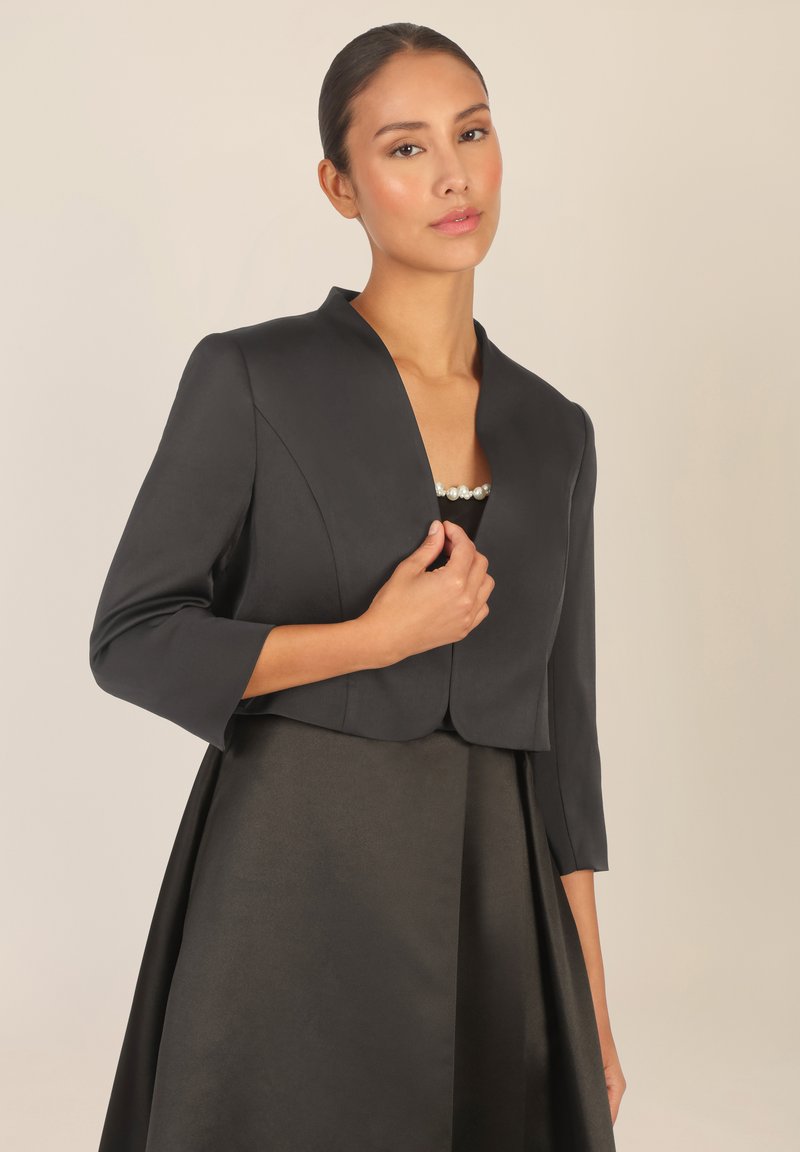 Mustat lyhyet takit, joissa on kolme neljäsosaa hihat, joissa on shaalikaulus ja sileä pinta, yhdistettynä tyylikkääseen, samasta kankaasta valmistettuun hameeseen.