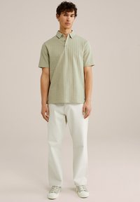 Polo de manga corta verde claro con un patrón texturizado y un cuello abotonado, combinado con pantalones blancos de pierna recta y zapatillas ligeras.