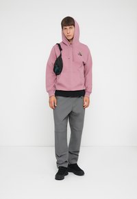 Pink hooded sweatshirt with logo, black underlayer, gray pants, and black shoes. Small black bag slung over shoulder. Simple, casual look.