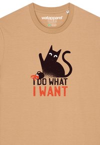 Beige katoenen t-shirt met een grafisch ontwerp van een zwarte kat met grote ogen en de tekst "IK DOE WAT IK WIL" in vette, rode en zwarte letters.