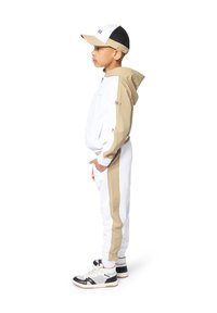 Completo sportivo composto da una giacca bianca e beige con zip, pantaloni bianchi abbinati e sneakers con accenti neri, accompagnato da un cappellino bianco e nero.