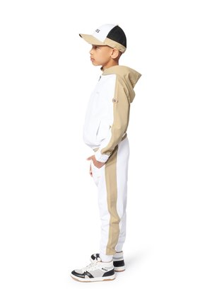 Vêtement de sport comprenant une veste zippée blanche et beige, un pantalon blanc assorti et des baskets avec des accents noirs, le tout accompagné d'une casquette blanche et noire.