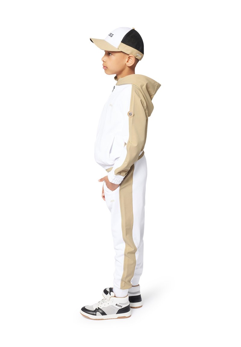 Completo sportivo composto da una giacca bianca e beige con zip, pantaloni bianchi abbinati e sneakers con accenti neri, accompagnato da un cappellino bianco e nero.