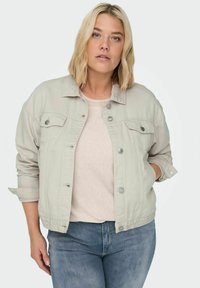 Chaqueta de mezclilla gris claro con cuello tipo camisa, botones a presión en el frente y bolsillos laterales. Lleva puesta una blusa beige con mangas enrolladas.