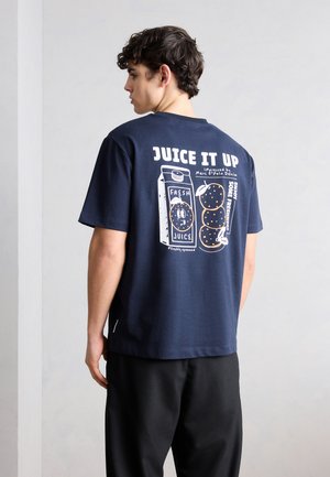 Mladý muž v tmavomodrom tričku s grafikou „JUICE IT UP“, ktorá zobrazuje pomaranče a kartón džúsu na zadnej strane, stojaci pred obyčajnou stenou.