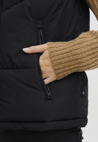 Svart puffv�äst med en strukturerad, ribbad beige tröjärm. Har sidofickor med dragkedja och en dragsko nederdel för justerbar passform.