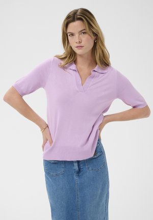 Ženska s svetlimi lasmi, oblečena v lilijasto polo majico s kratkimi rokavi in modro jeans krilo, stoji z rokami na bokih pred belim ozadjem.