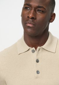 Beige Strick-Polohemd mit strukturiertem Muster, ausgestattet mit einem Kragen und drei grauen Knöpfen auf der Vorderseite.