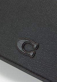 Sort tekstureret læderpung med en glat, blank metalaccent formet som et "C" på forsiden. Har et fint, detaljeret mønster hele vejen igennem.