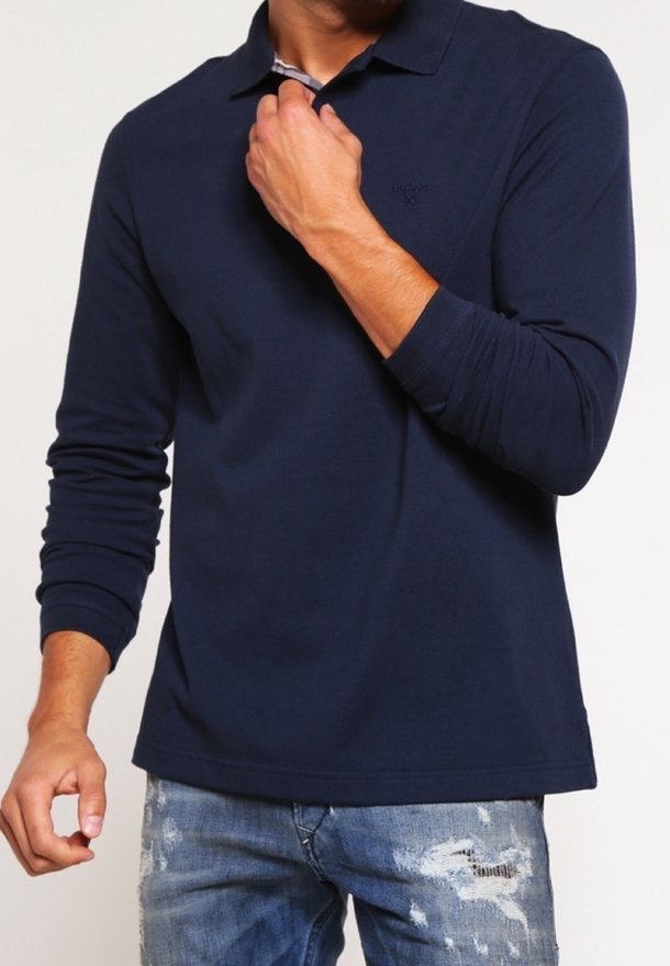 Homme portant un polo bleu marine à manches longues et un jean bleu déchiré ajustant le col de la chemise d'une main.