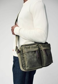 Sac messager en toile vert olive avec deux poches avant, accents en cuir marron, sangle réglable et tissu doux et texturé.