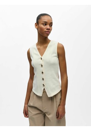 Frau trägt eine ärmellose cremefarbene Weste mit Knöpfen und beige hoch taillierte Hose, blickt seitlich vor einem schlichten weißen Hintergrund.
