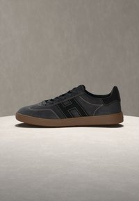 Baskets noires profil bas avec semelle marron et logo noir "H" sur le côté, exposées sur une surface ronde claire sur fond beige uni.