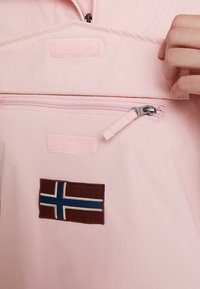 Różowa tkanina kurtki z kieszenią na zamek błyskawiczny i zapięciem na rzep. Posiada brązową łatę z wzorem niebiesko-białej flagi.