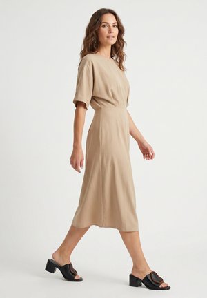 Kvinde går mod højre iført en beige midi kjole med korte ærmer og sorte åbne mulehjem med hæl mod en ens hvid baggrund.