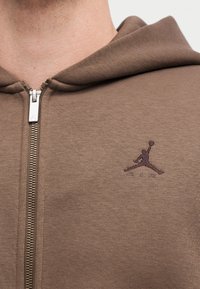 Gros plan d'une personne portant un sweat à capuche marron zippé avec un petit logo Jumpman brodé brun foncé sur la poitrine.