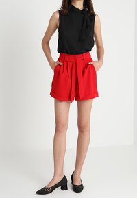 Black sleeveless top with a bow detail; red high-waisted shorts with a tied belt. Black woven pumps complete the outfit.