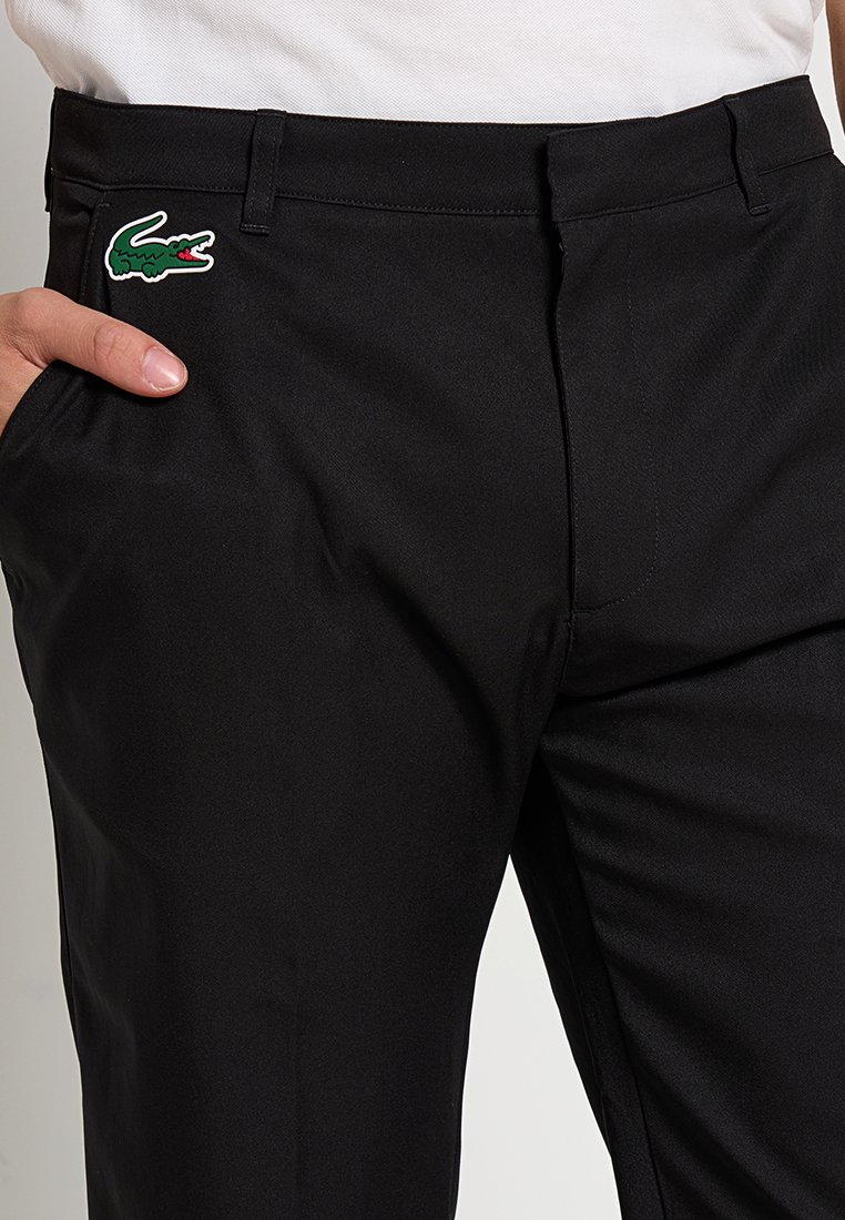 Pantalon noir avec une texture lisse, arborant un logo de crocodile brodé vert et blanc sur la zone de la poche gauche. Design ajusté.