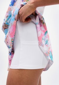 Χέρι που σηκώνει πολύχρωμη φούστα για να αποκαλύψει λευκά στενά σορτς με πλαϊνή τσέπη στον μηρό ενός ατόμου.