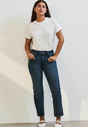 Witte katoenen t-shirt, donkerblauwe hoge taille jeans, en witte schoenen met zwarte stippen. Model poseert met handen in de zakken tegen een muur.