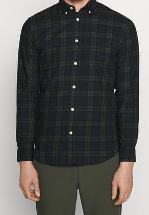 Mann trägt ein schwarz-grünes kariertes Hemd mit langen Ärmeln und grüne Hosen, steht mit entspannten Armen an den Seiten.