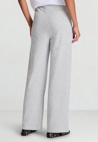 Graue Sweatpants mit weitem Bein und glatter Textur, elastischem Bund und schwarzen Schuhen mit silbernen Ösen am Saum.