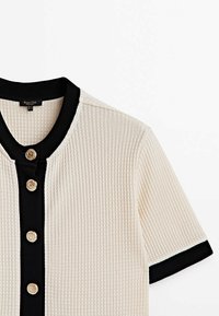 Haut en maille crème à manches courtes avec bordure noire au col, à la patte de boutonnage et aux bords des manches, orné de quatre boutons décoratifs dorés.