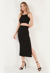 Un crop top noir et une jupe taille haute avec une fente haute sur la cuisse. Tissu lisse, design ajusté, et associé à des sandales noires à talons hauts.