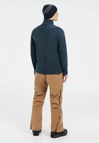 Laivyno mėlynos spalvos polarinis džemperis su aukštu apykakle, derinamas su smėlio spalvos kargo kelnėmis, turinčiomis užtrauktukais uždaromas kišenes ir šoninius skilimus apačioje. Juodi batai.