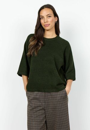 Vrouw met lang donker haar, die een donkergroene korte mouw trui en geruite broek draagt, staand met de handen in de zakken tegen een witte achtergrond.