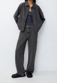 Giacca in denim grigia oversize con chiusura a bottoni, abbinata a una canotta aderente a righe e pantaloni grigi larghi. Scarpe nere slip-on.